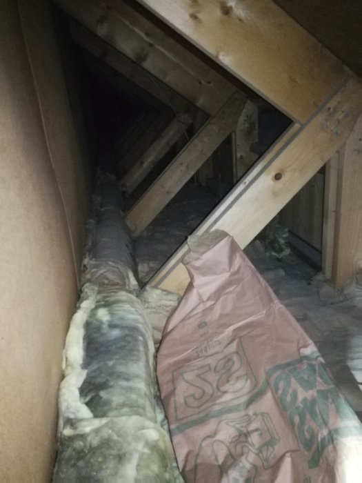 Kattvind med synliga träbjälkar och oregelbundet placerad isolering, vissa delar blottade.
