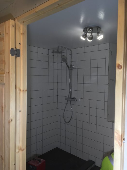 Ett nyligen installerat takduschsystem och taklampa i en dusch med vita kakelväggar och mörkt golv, sett genom en öppen dörr.