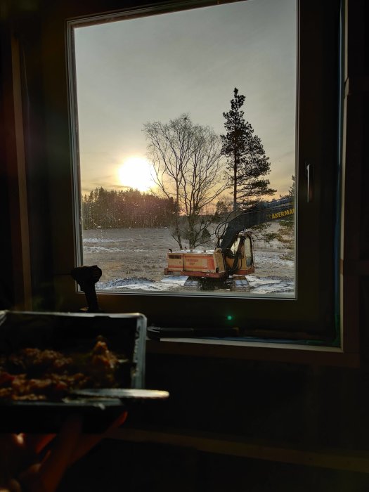 Solnedgång utanför ett fönster med utsikt över ett vinterlandskap och en grävmaskin, förgrund visar en hand som håller en lunchlåda.