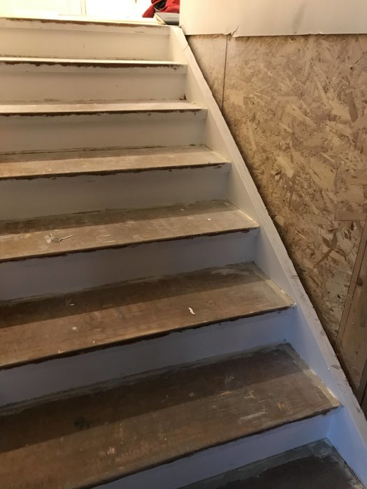 Ommålade trätrappsteg med delvis borttagna sidostycken nära en vägg av OSB-skivor, i behov av förstärkning.