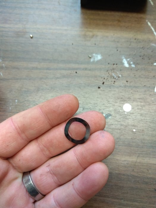Hand håller en misstänkt sliten metallring, troligtvis en del av en motor, mot en träbänk.