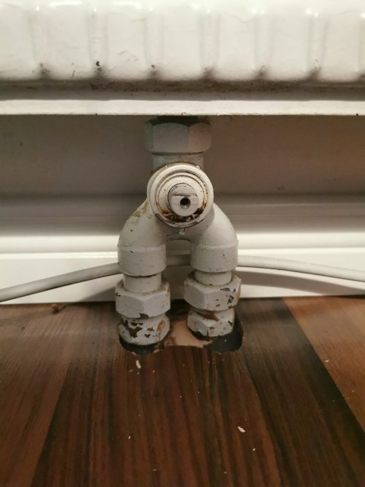 Skadad och rostig radiatorventil med skalad färg på trägolv framför vit element.