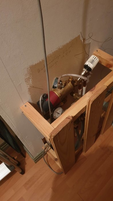 Värmepumpens installation med rörelser, kopplingar och en manometer inramad av trästöttor mot en grå vägg.