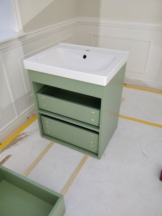 Grönt tvättställsskåp med två utdragna lådor och en vit handfat ovanpå, i ett rum under renovering.