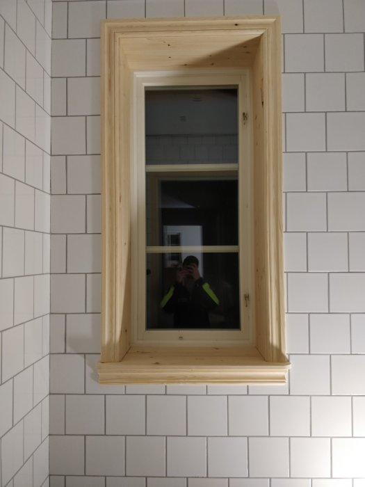 Träramad spegel på vit kaklad vägg med reflexion av person som fotograferar.