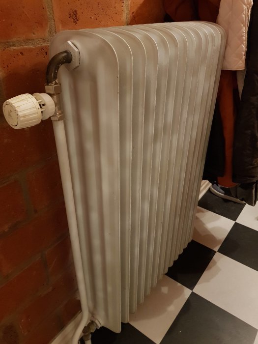 Gammal vit stående radiator med termostatventil mot tegelvägg, vid ett schackrutigt golv.