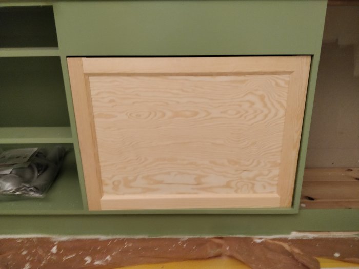 Hemsnickrad kökslucka med ram av planhyvlat trä och inlägg av furuplywood mot grönmålad skåpstomme.