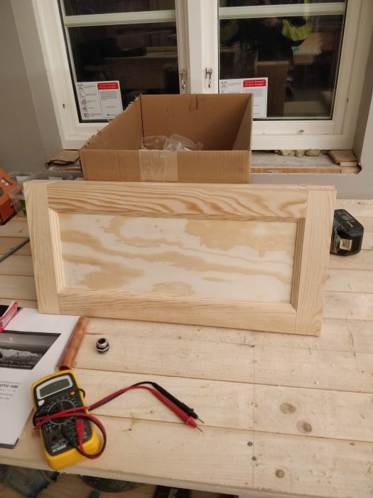 Hemsnickrad kökslucka av furu med profilerade ramdelar och plywood spegel på ett byggbord, verktyg i förgrunden.