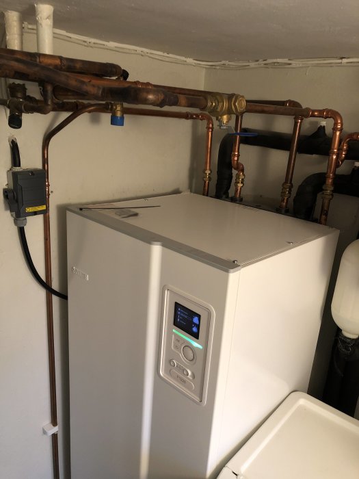 Värmepump och kopparledningar i pannrum, med kontrollpanel och diverse ventiler.