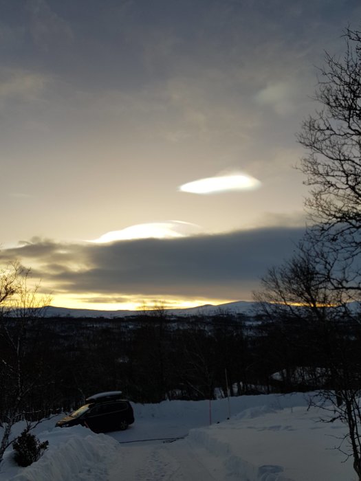 Vintersolnedgång med linsmoln över snötäckta landskap och en bil.