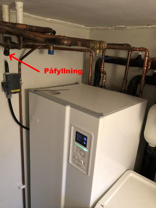 Värmepump i pannrum med kopparledningar och röd pil som markerar påfyllningsventil.