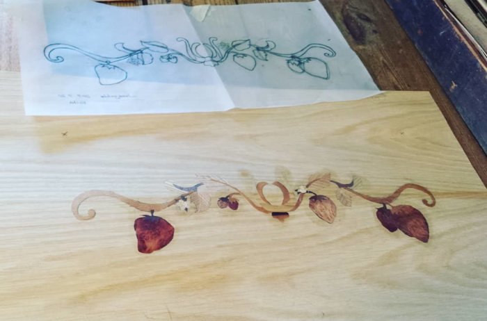 Intarsiaarbete som visar en blomranka i trä bredvid dess skiss på papper.