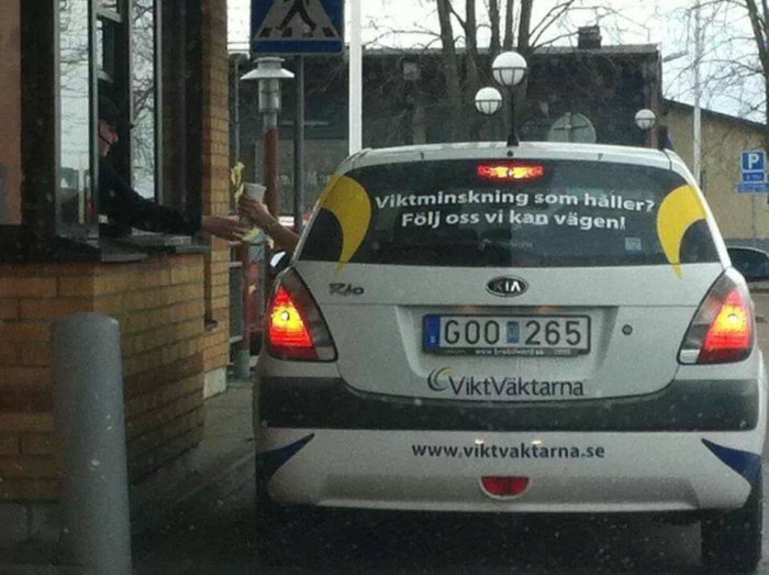 Bil vid drive-thru vidtar emot matbeställning, med reklam för viktminskning på bakluckan.