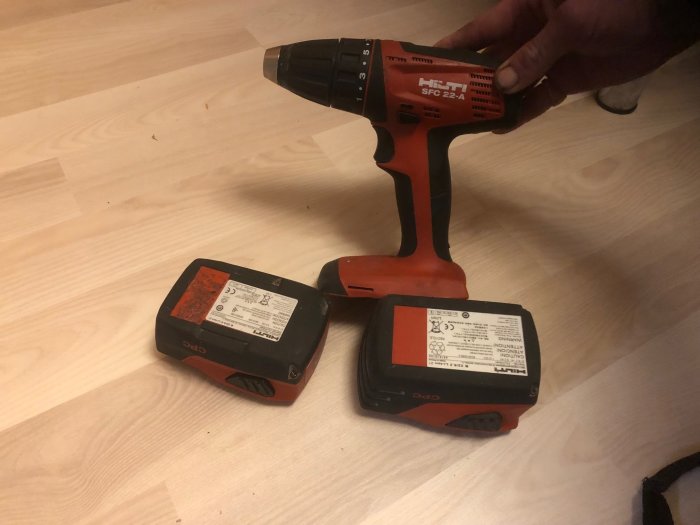 En hand håller en orange och svart batteridriven skruvdragare bredvid två batterier på ett trägolv.