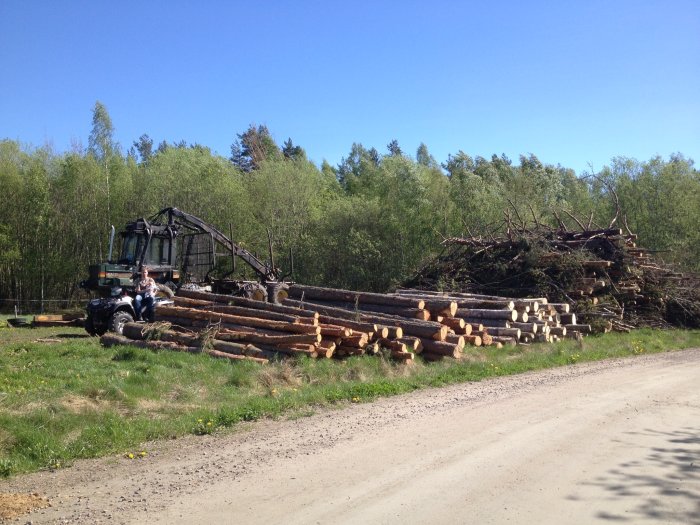 Skogsmaskin som lastar timmer vid en röjd tomt, högar med virke och ris i bakgrunden.