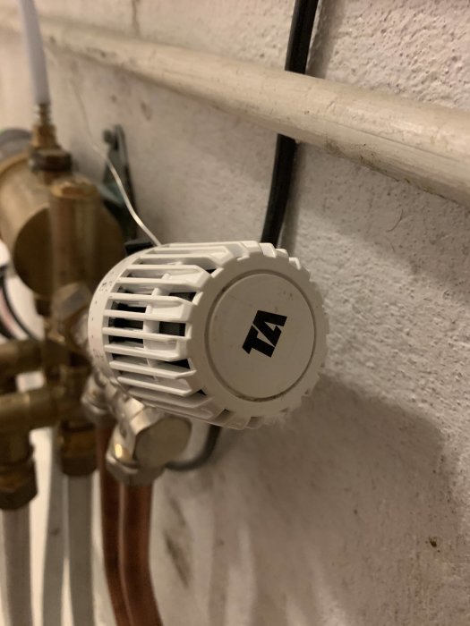 Termostatventil på radiator med synliga rör och ventil mot en vit vägg.