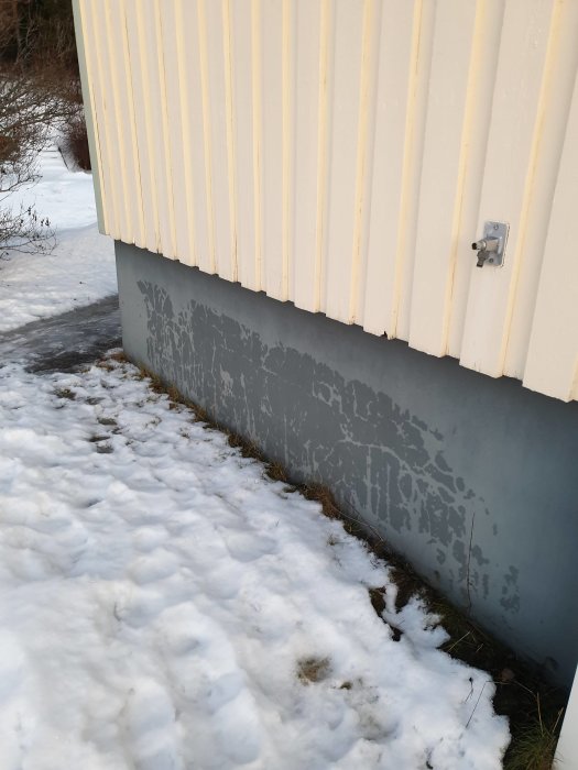 Husgrund med fuktmärken på betongvägg under gul panel, omgiven av snö och gräs.
