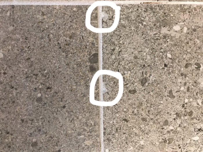 Närbild på betongyta med små urgröpningar markerade med vita cirklar vid kanten av kakelplattor.