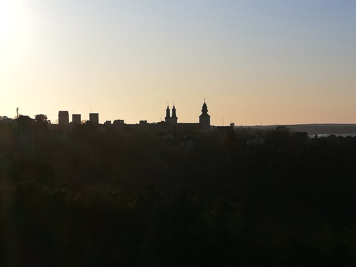 Stadsiluett vid solnedgång med kyrktorn och byggnader i motljus och träd i förgrunden.