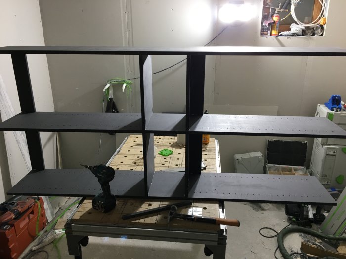 Nybyggd svart bokhylla med synliga domino-fogar på en arbetsbänk i ett verkstadsutrymme.