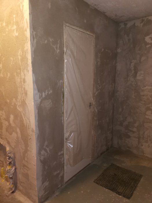 Oputsade väggar i en källare med ojämn och fläckig puts, en ingång dörr täckt med plast och en matta på golvet.