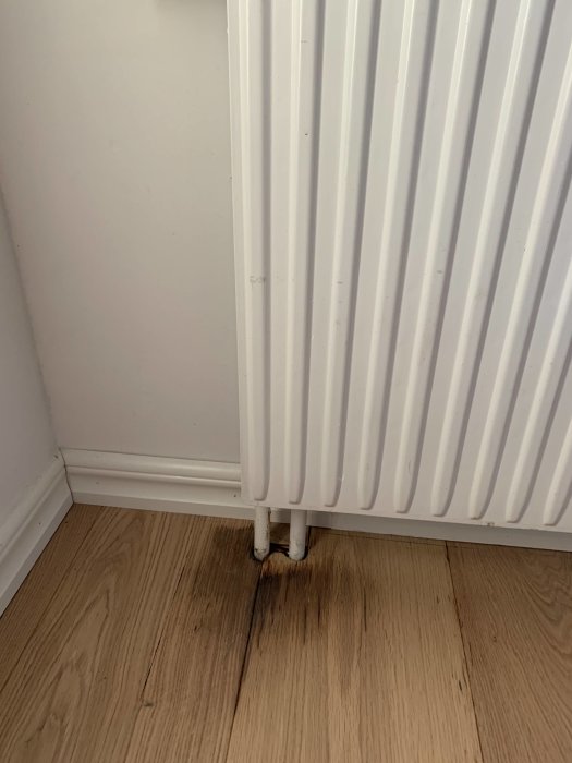 Missfärgat trägolv vid vitt radiatorrör och fotlist, potentiell vattenskada.