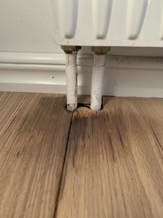 Missfärgat trägolv vid elementrör, tecken på möjlig vattenskada från röret eller blöta skor.