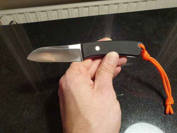 Hand håller en kniv utan egg med svart G10-skaft och orange snodd.