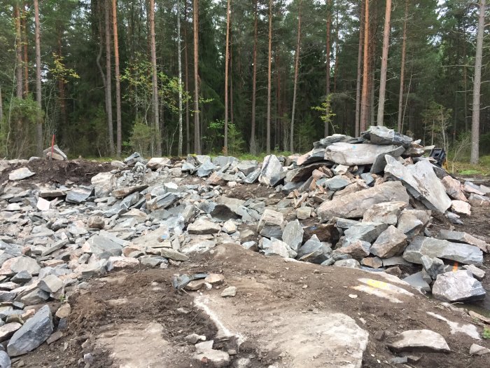 Sprängningsresultat med stenar och bergstycken på en byggplats i skogsmiljö, grävmaskin i bakgrunden.