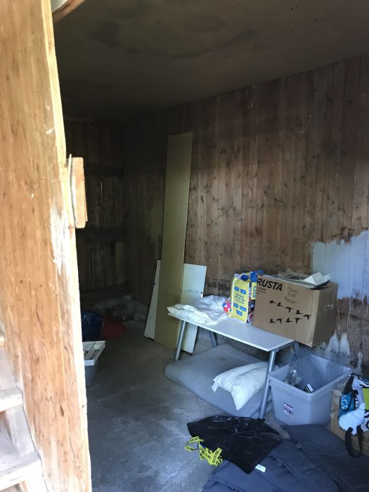 Oisolerat rum med träläktväggar, betonggolv, en öppen dörr och diverse föremål utspridda.