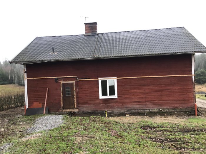 Rött trähus med mörkt tak och skorsten, delvis renoverat, i lantlig miljö.