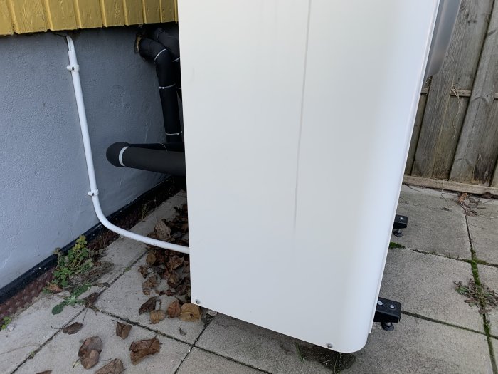 Utedel av en luft-värmepump monterad på plattor med synliga rör och kablar intill en husvägg.