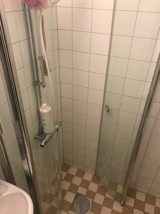 Ett mindre badrums hörndusch med öppen dörr, vet vita kakelväggar och beiget klinkergolv.