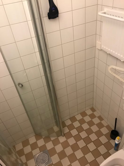 Mindre badrum med hörndusch, vitt kakel, beige-brunt golv, och vägghängd toalett till höger.