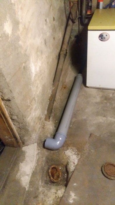 Grått WC-rör med 90 graders böj anslutet till gammal gjutjärnsmuff i betonggolv i källare.