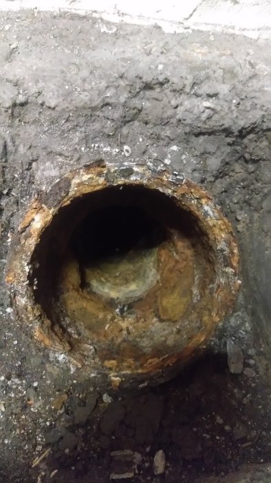 Gammal rostig gjutjärnsmuff i betonggolv, potentiell renslucka, med mörkt avloppshål.