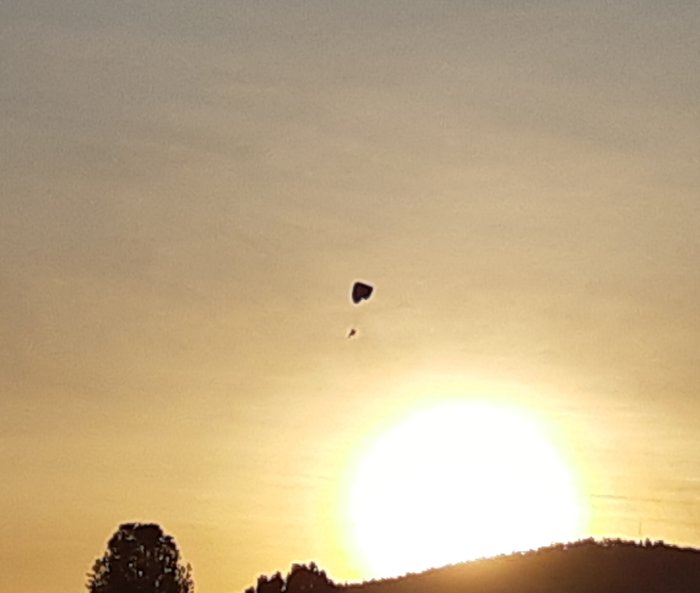 Luftballong flyger mot en solnedgång med träd och kullar i silhuett.