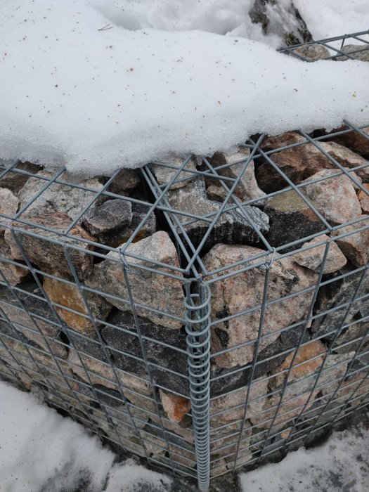 Stenar i gabionvägg med snötäcke och synlig ståltrådsförstärkning.