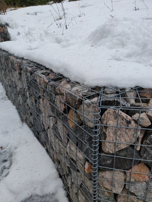 Gabionmur av sten och ståltråd delvis täckt av snö.