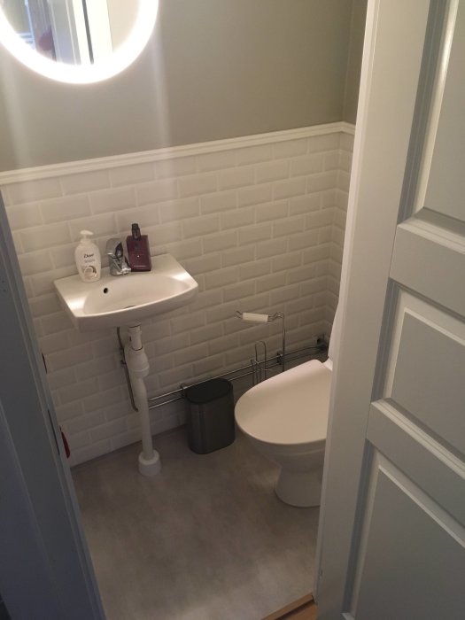 Renoverat badrum med vita kakelväggar, handfat, toalett och grått golv, reflektion av en ljusring speglas i spegel.