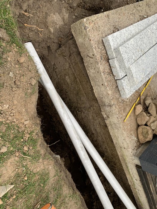 Grävd grävning bredvid garage med vit rörledning och exponerad isolering vid betongvägg.