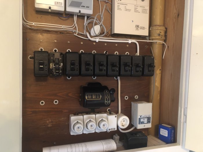 Gammal elcentral med flera svarta automatsäkringar och ett öppet säkringsskåp, på en trävägg.