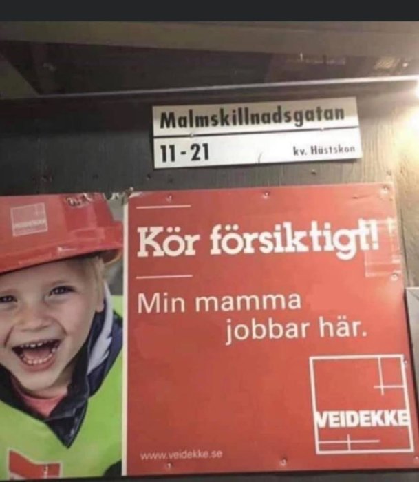 Skylt med texten "Kör försiktigt! Min mamma jobbar här." och leende barn i bygghjälm.