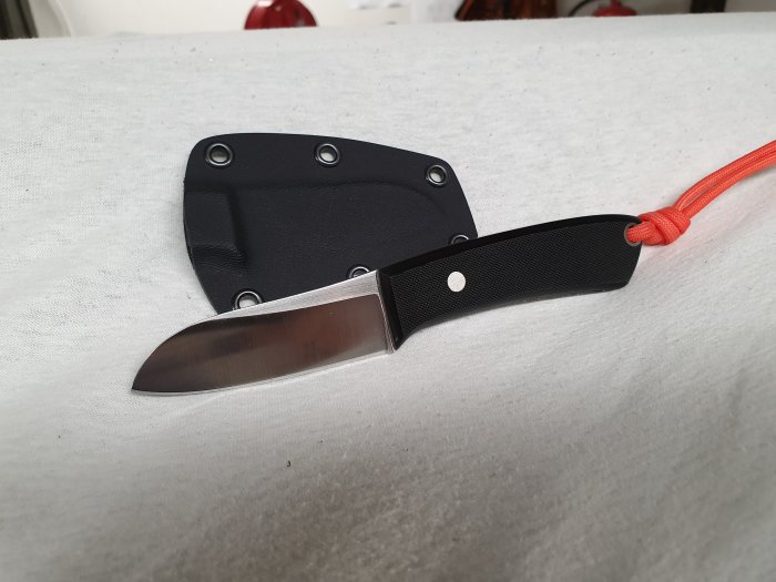 Kniv med svart handtag och orange snodd, bredvid svart knivslida, på ljus bakgrund.