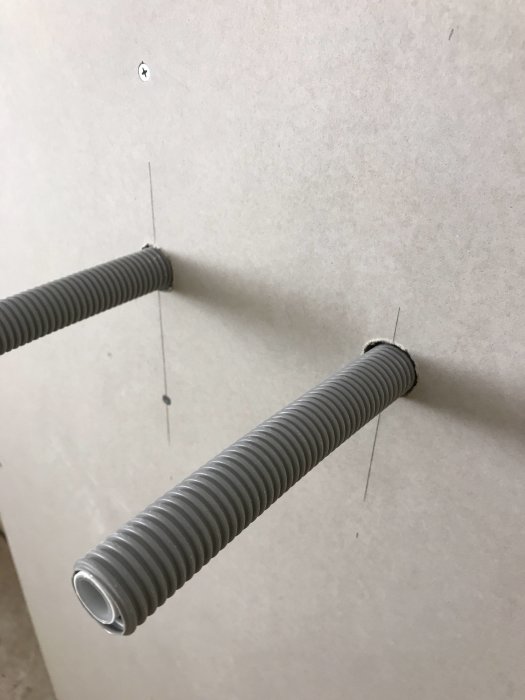 Två gråa flexibla rör-i-rörsystem som sticker ut ur en vit plywoodvägg under renovering av badrum.