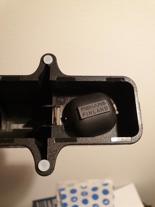 Svart plastdel med texten "FISKARS FINLAND" som en del av en byggprodukt, utan lapp, på en otydlig bakgrund.