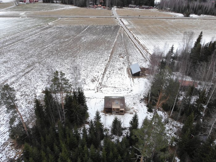 Drönarbild över en byggplats med halvfärdigt hus omgivet av snötäckt skog och åker.