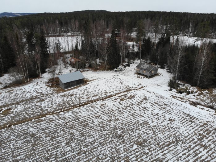 Flygbild på en tomt med snötäckt mark, en byggnad under konstruktion och ett skogsparti.