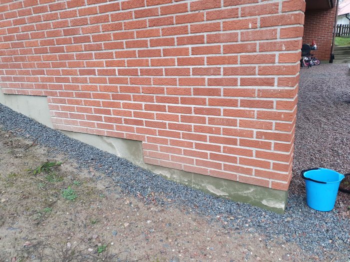 Tegelfasad på suterränghus med fuktfläckar vid grunden och husknuten, grus och hink på marken.