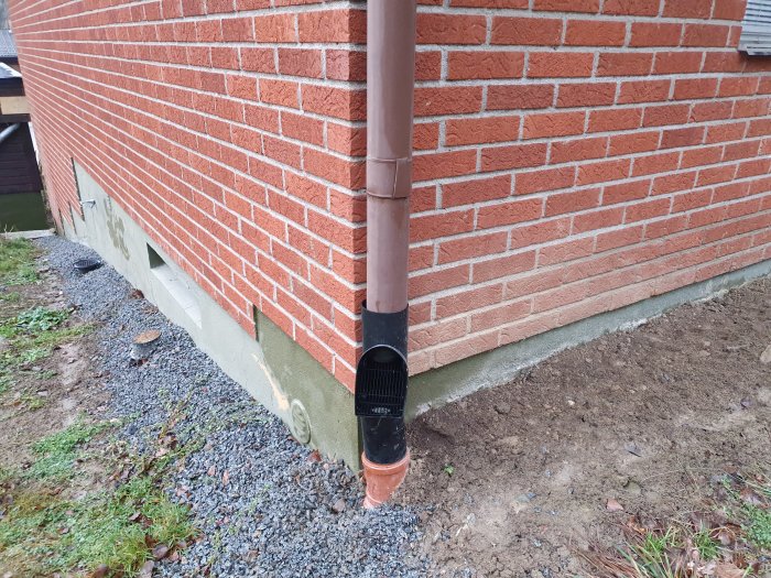 Fuktfläckar på betonggrund och tegelvägg vid husknut med nedrör och grus.